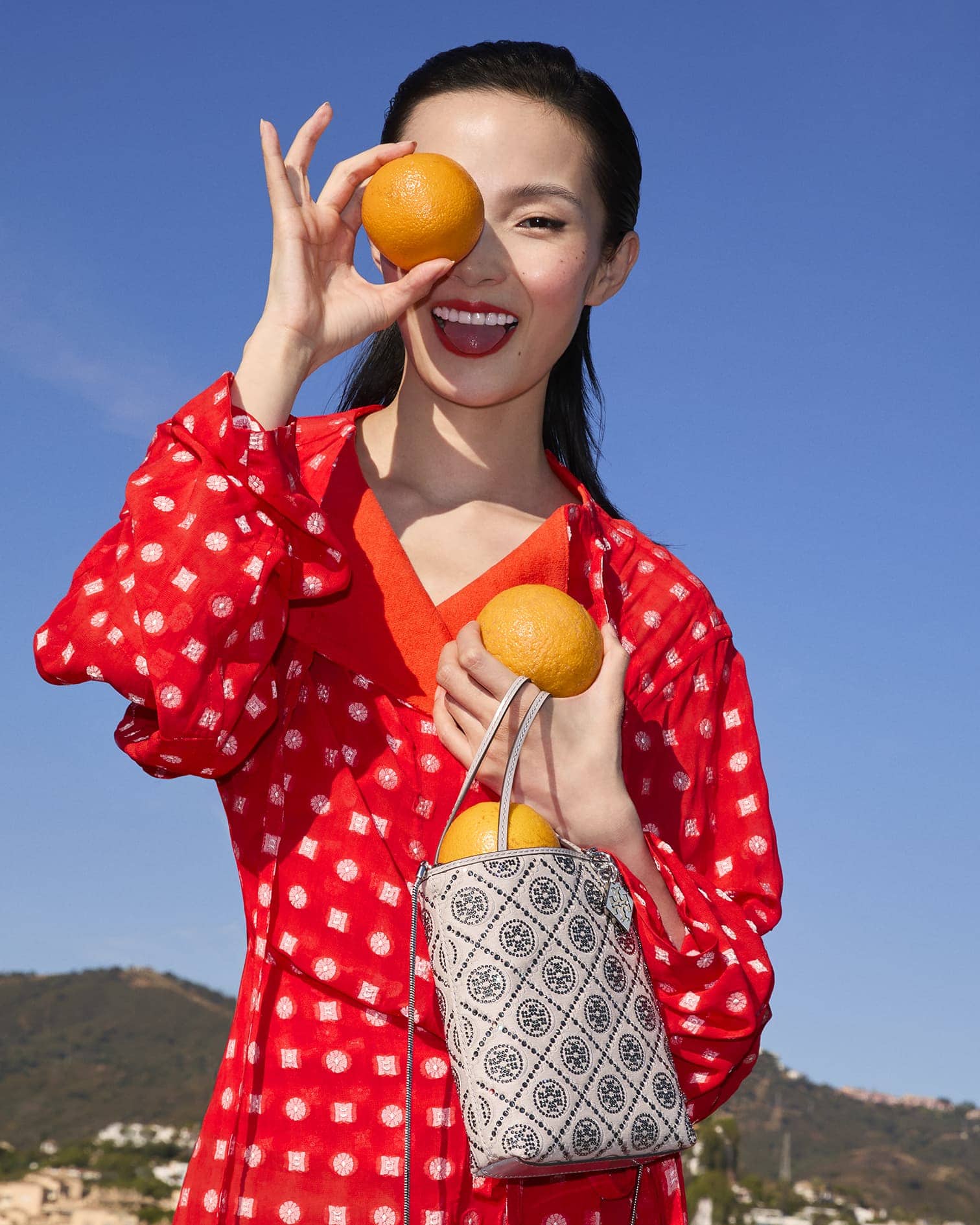 Xiao Wen Ju holding oranges in front of her eye wearing a red dress and patterned handbag under a clear sky