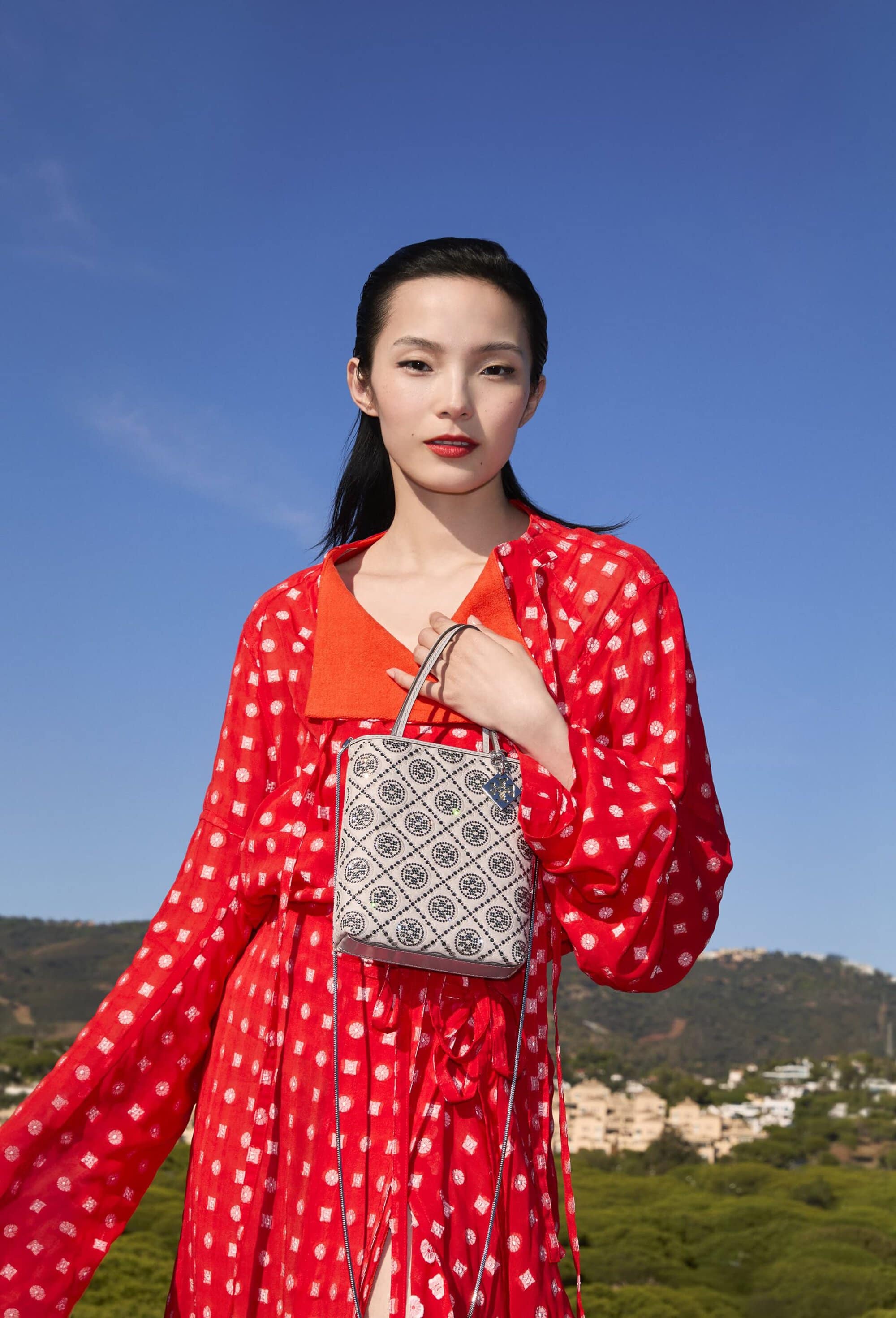 Model Xiao Wen Ju in a red patterned dress holding a patterned handbag against a clear blue sky and hilly landscape