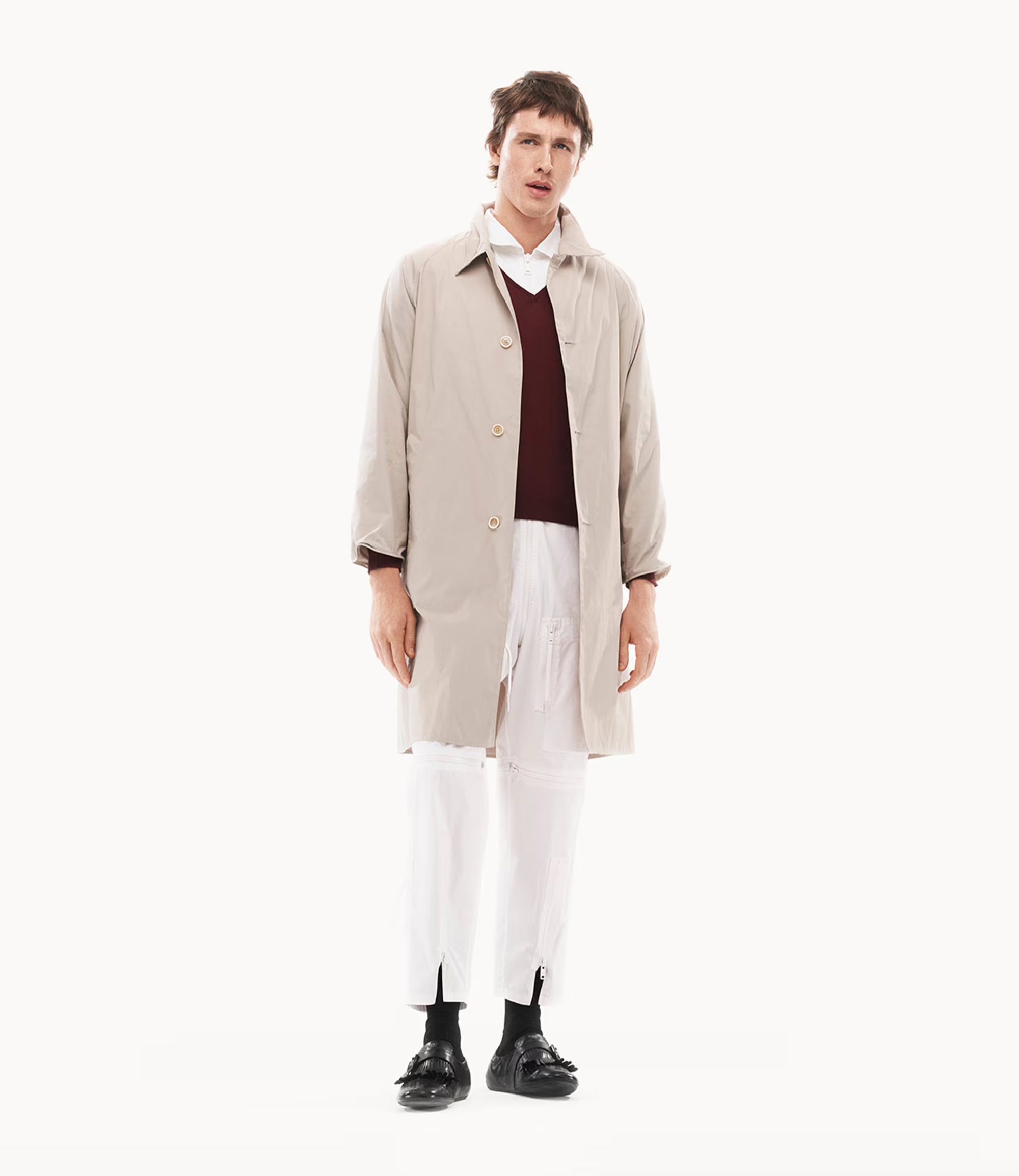 Harris Dickinson wearing a beige coat over a burgundy sweater and white trousers against a white seamless background