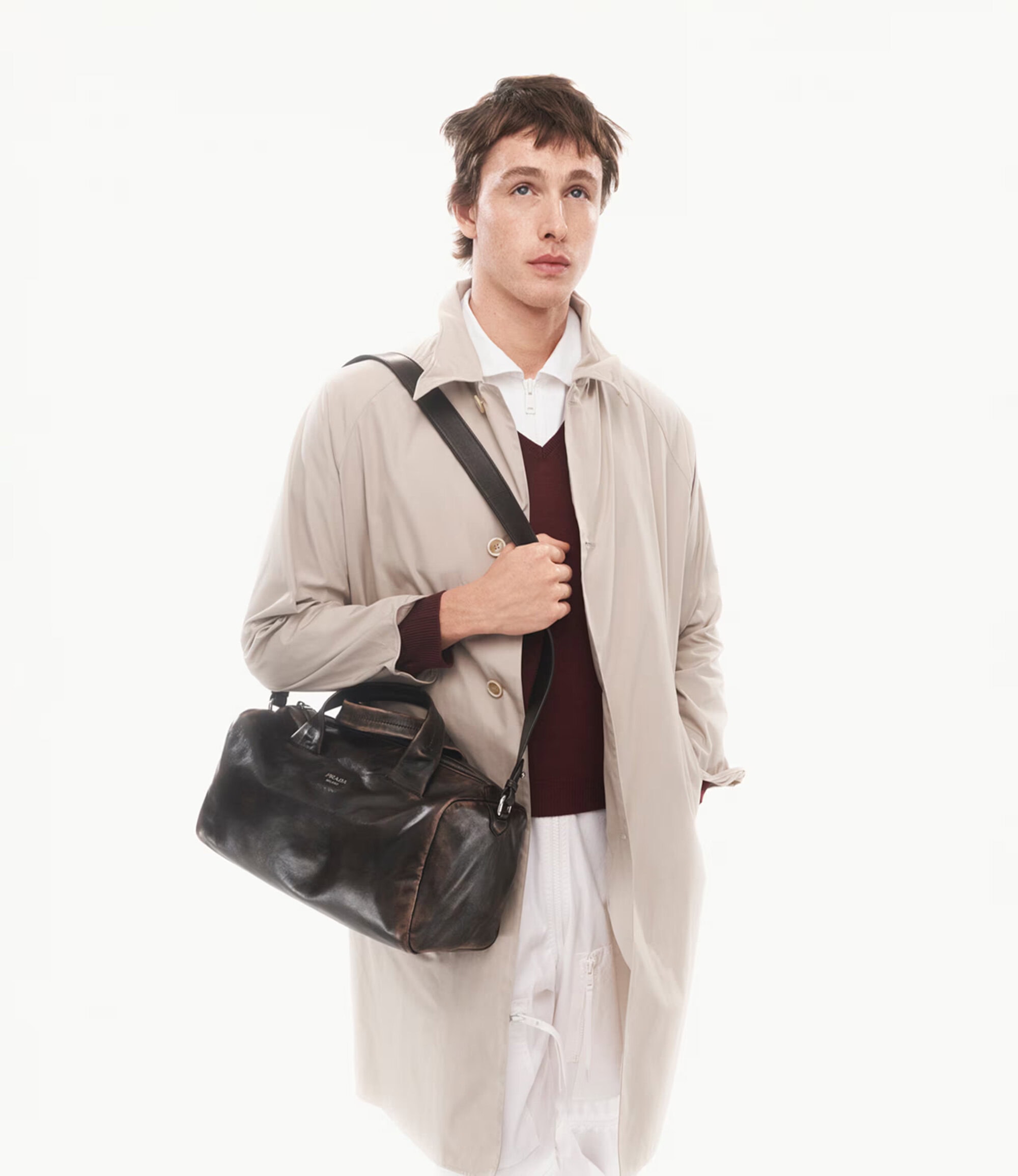 Harris Dickinson wearing beige trench coat and carrying a dark leather duffel bag against a white background