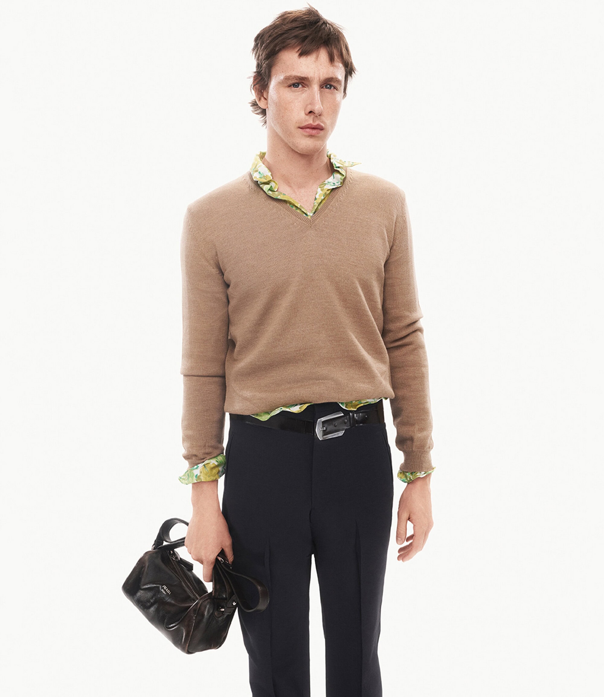 Harris Dickinson in a tan v-neck sweater and dark trousers holding a handbag against a white background for Prada.