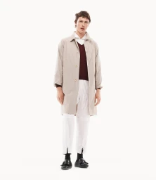 Harris Dickinson wearing a beige coat over a burgundy sweater and white trousers against a white seamless background
