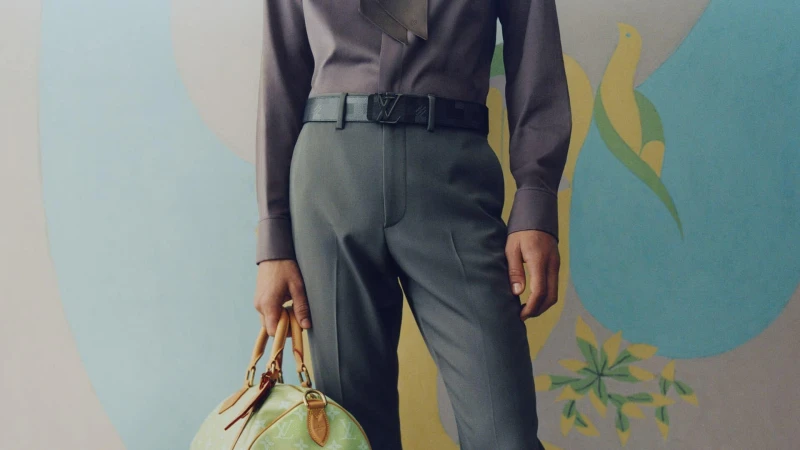 Close-up of a man wearing grey trousers, a purple shirt with a necktie, a patterned belt, and holding a light green monogram handbag.