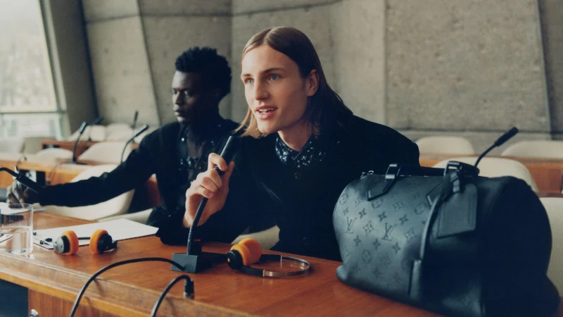 Two men seated at a wooden conference table with microphones and headphones, featuring a black duffel bag.