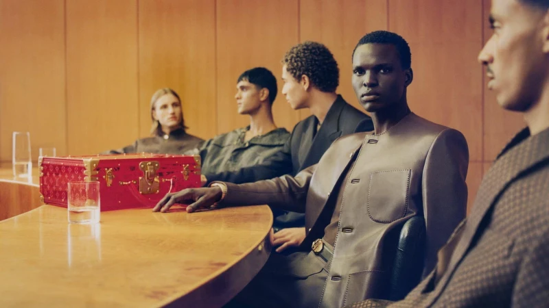 Five men seated around a polished wooden boardroom table with a bright red trunk case under warm lighting for Louis Vuitton Men's Spring 2025.