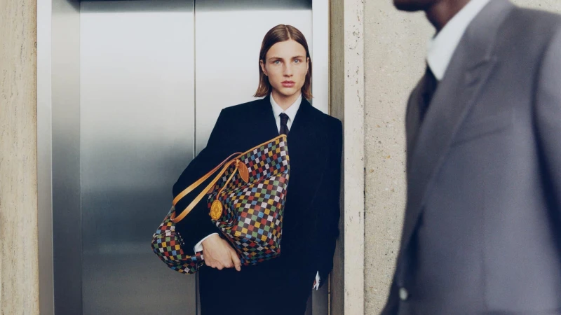 Model in dark suit holding a colorful checked tote bag standing in front of elevator doors