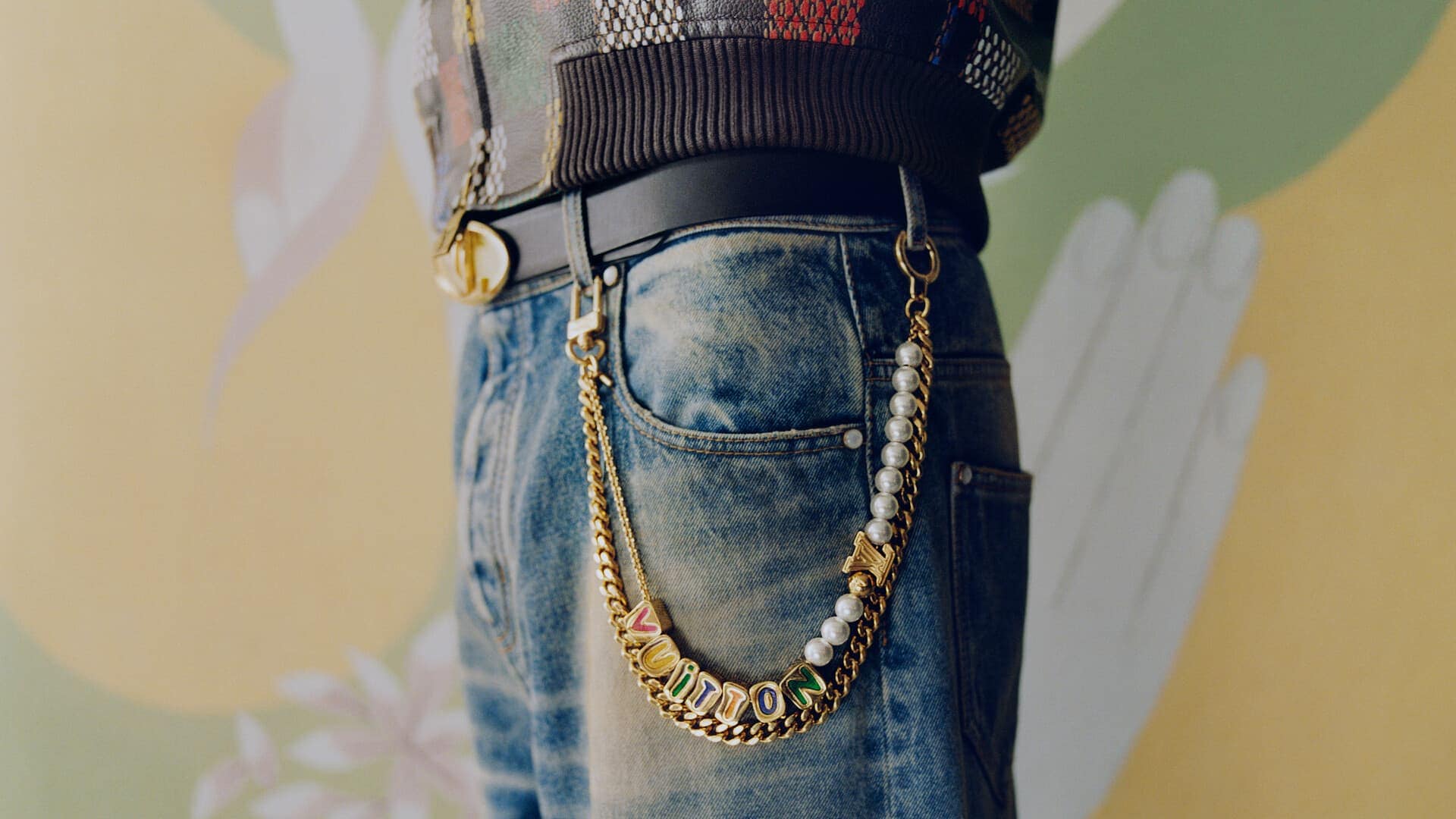 Close-up of denim jeans and a patterned jacket with a gold chain strap and pearl charm accessory for Louis Vuitton.