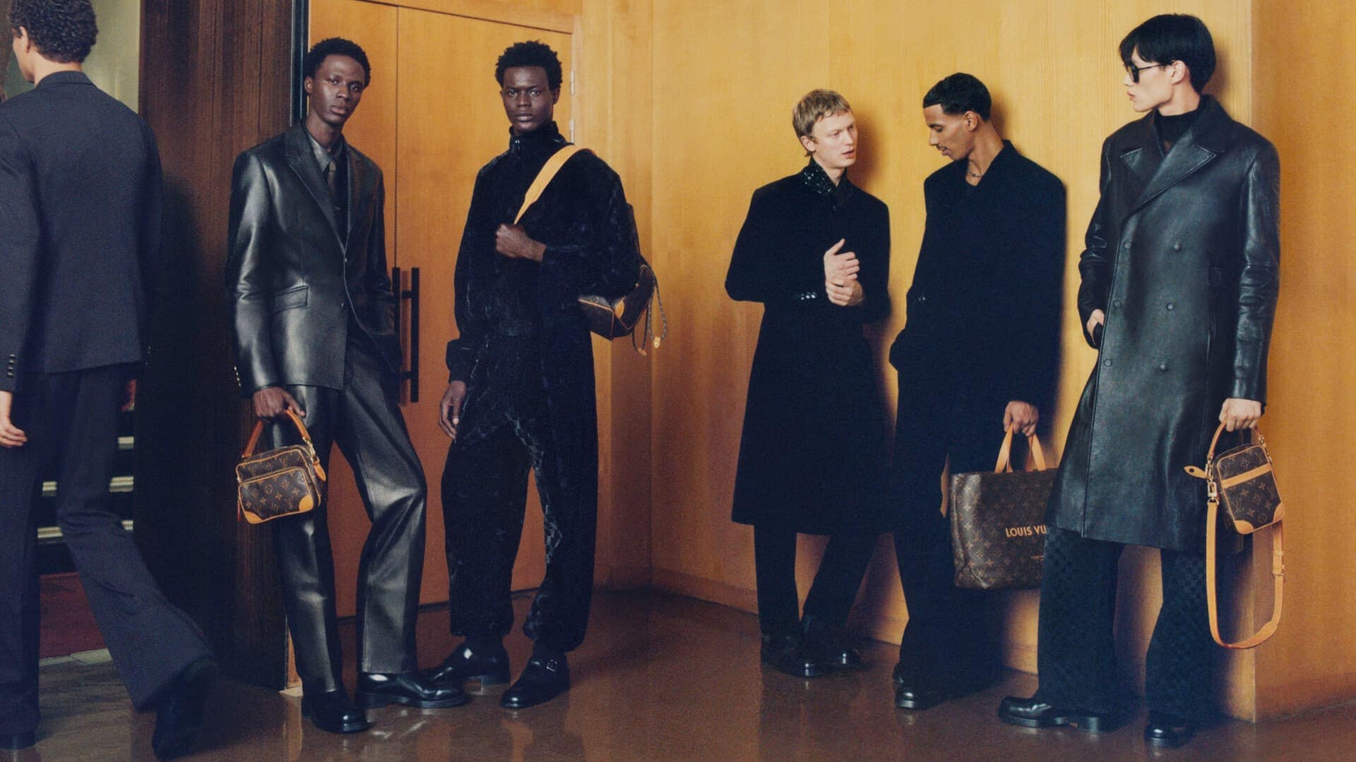 Group of six male models in black attire holding monogram handbags standing in a hotel lobby with wooden walls