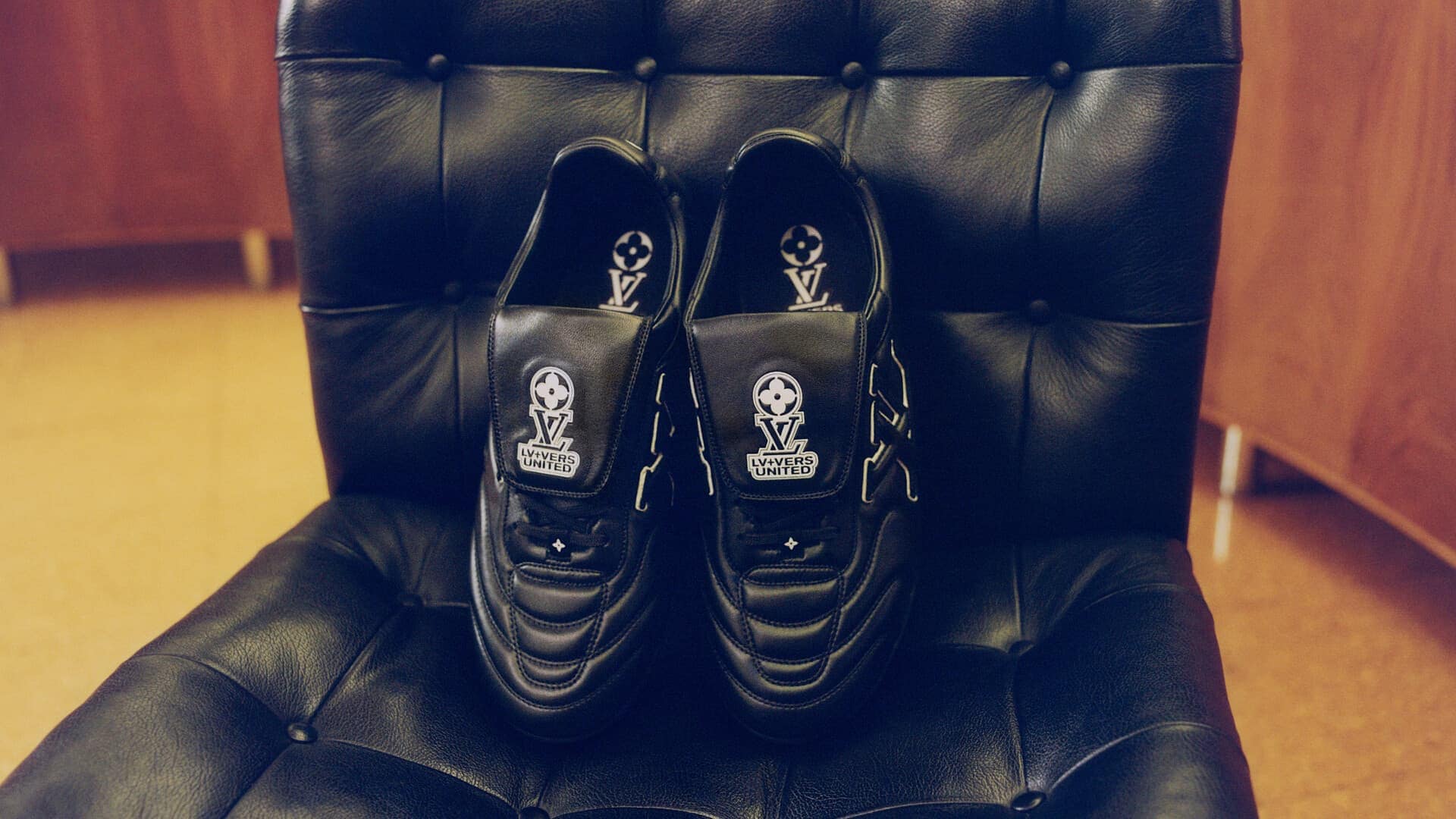 A pair of black leather sneakers with white logo branding placed on a tufted black leather armchair.