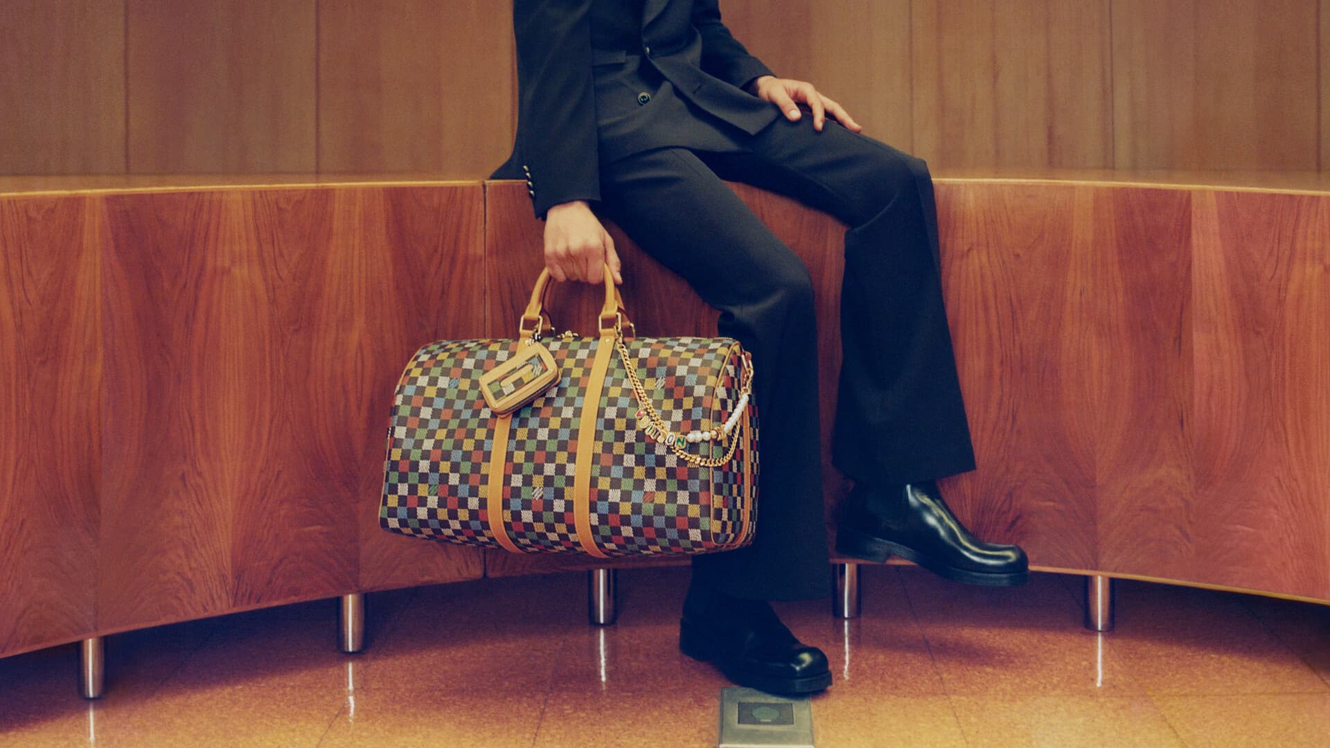 Man seated on wood credenza holding a checkered leather travel duffel bag for Louis Vuitton