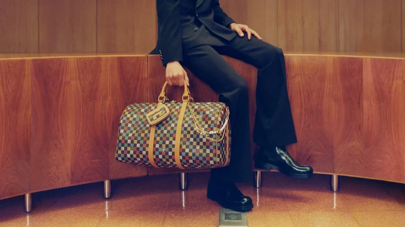 Man seated on wood credenza holding a checkered leather travel duffel bag for Louis Vuitton