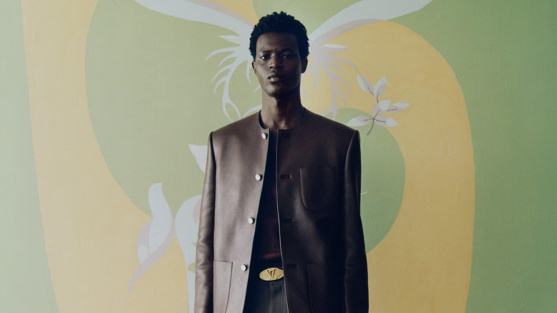A man wearing a brown leather jacket and belt with gold buckle against a green and yellow painted mural background