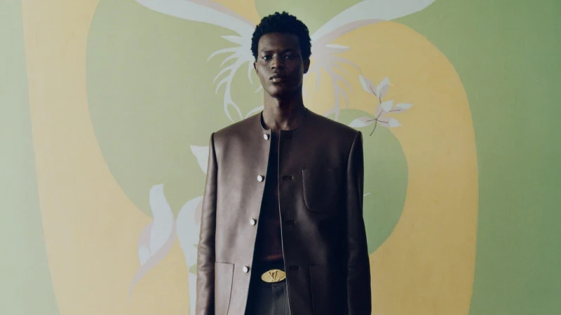 A man wearing a brown leather jacket and belt with gold buckle against a green and yellow painted mural background