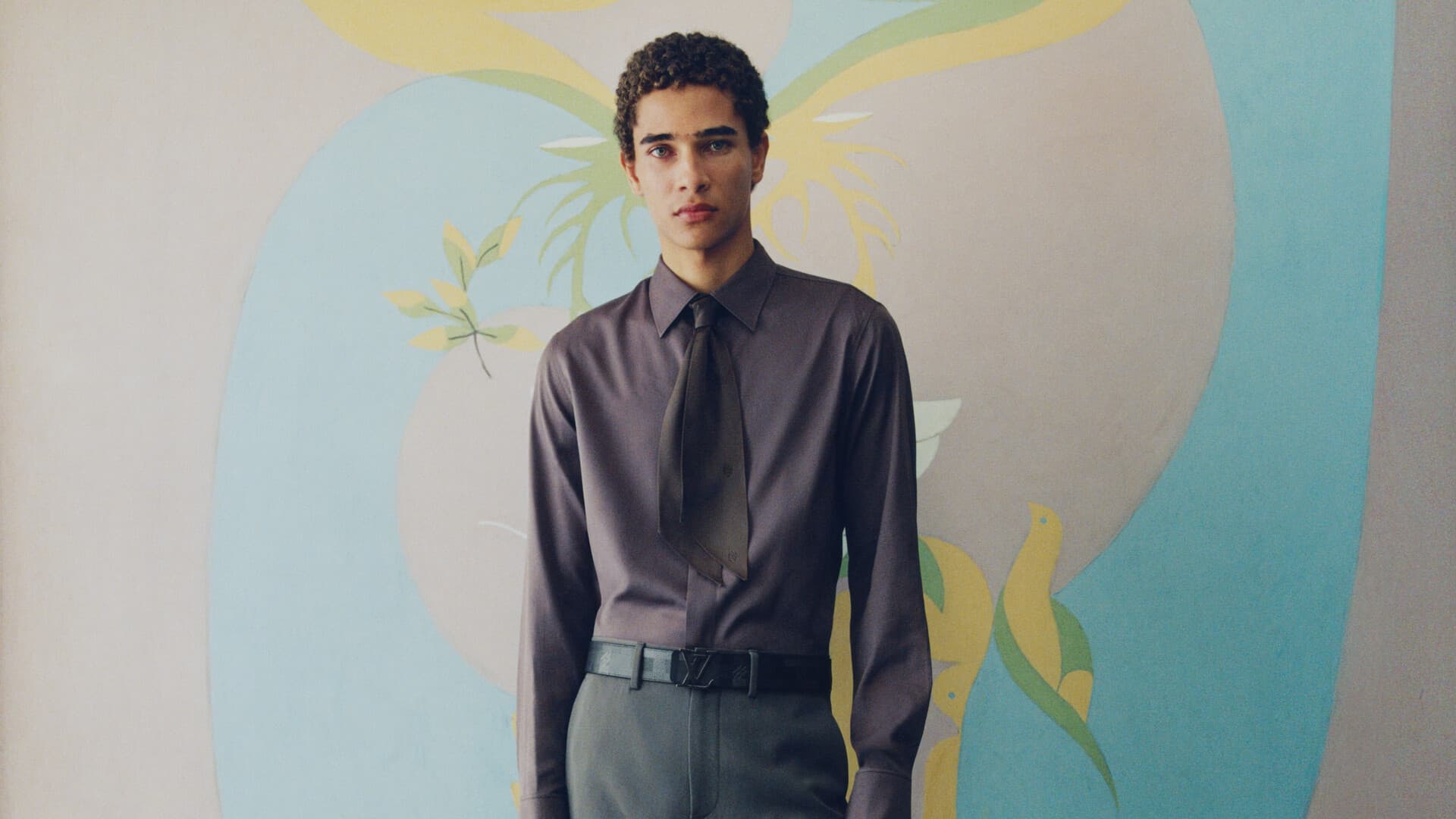 A young man wearing a grey shirt and tie standing against an ornate painted mural background for Louis Vuitton.