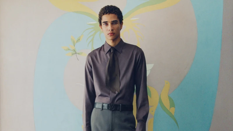 A young man wearing a grey shirt and tie standing against an ornate painted mural background for Louis Vuitton.