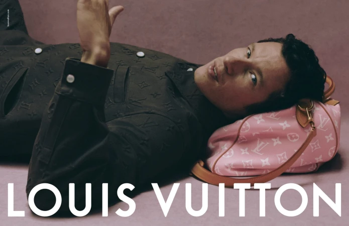 Man wearing a dark embossed jacket reclining on a pink textured background using a pink monogram duffle bag as a pillow