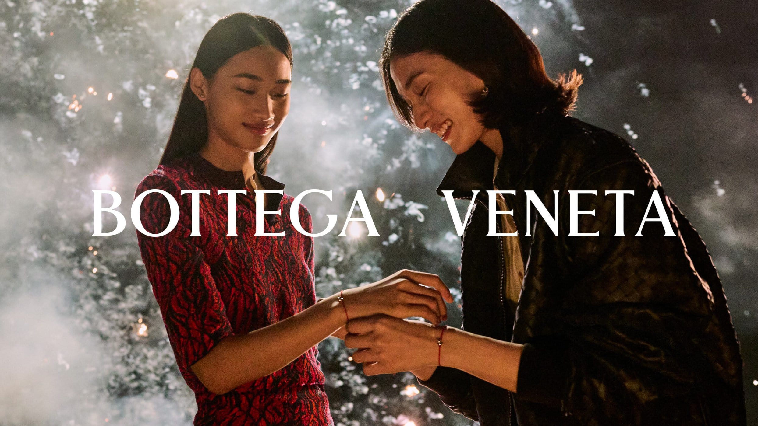 Two models tying a red string bracelet outdoors at night with sparkling bokeh lights in the background for Bottega Veneta