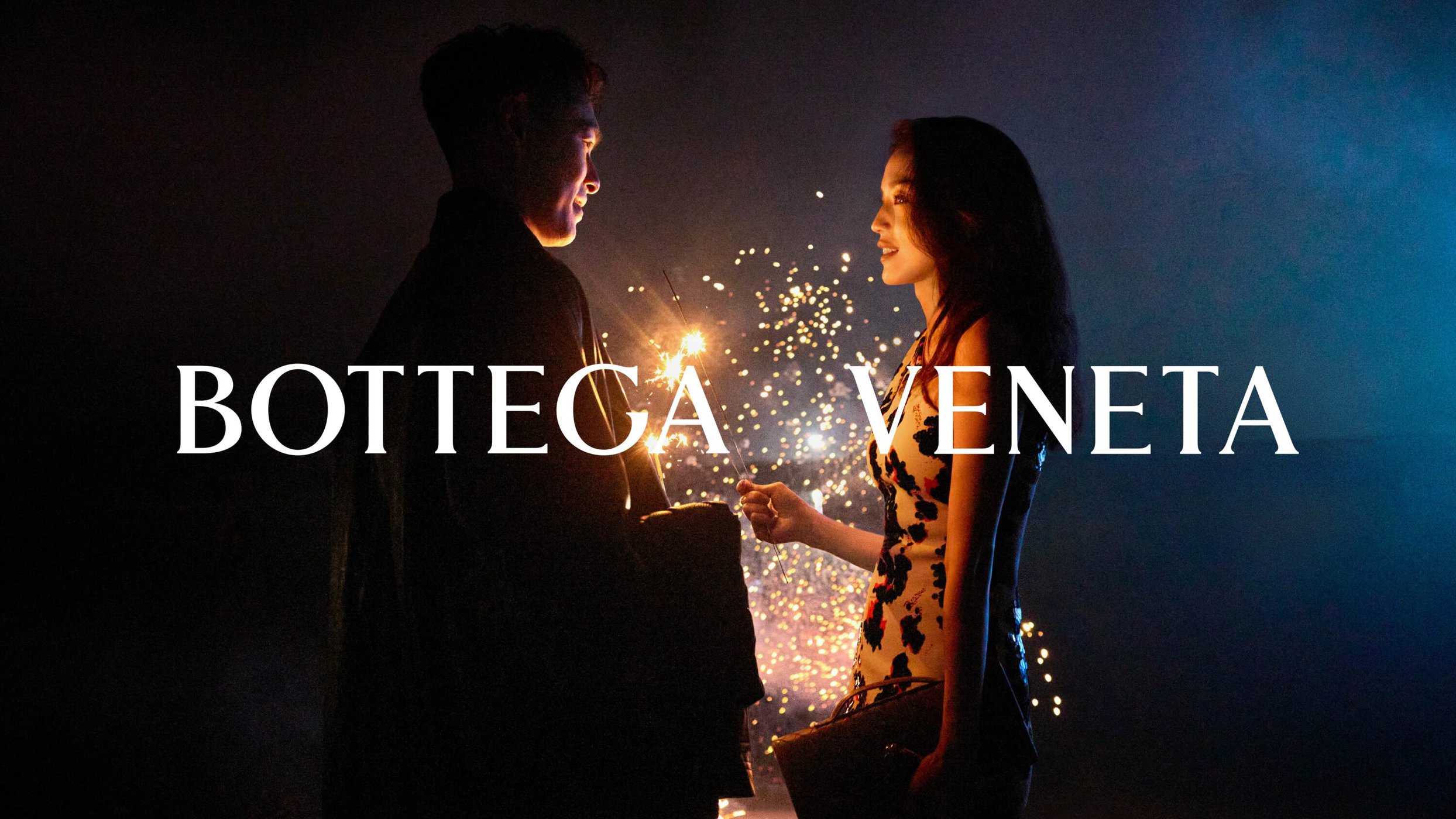 Man and woman holding sparklers outdoors at night with Bottega Veneta campaign text