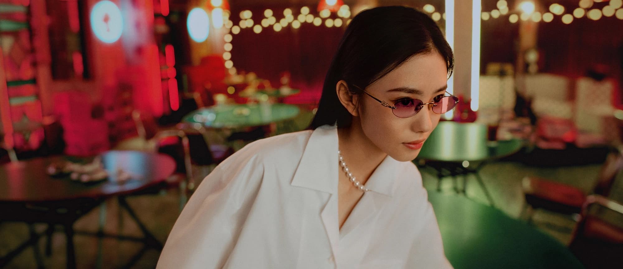 A young woman wearing tinted sunglasses and a white collared shirt in a warmly lit interior space with tables and glowing string lights.