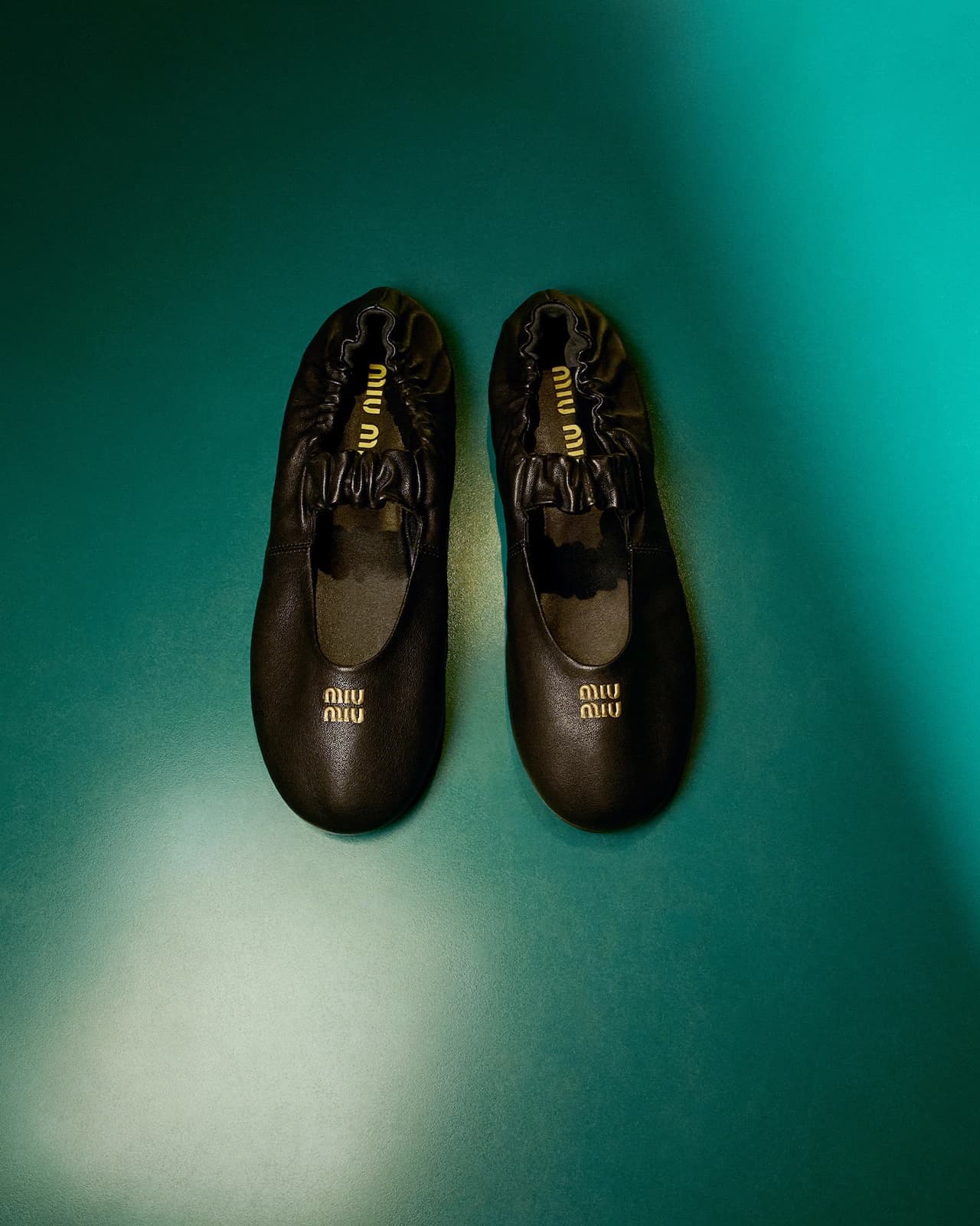 A pair of black leather ballet flats with gold branding presented vertically against a teal background.