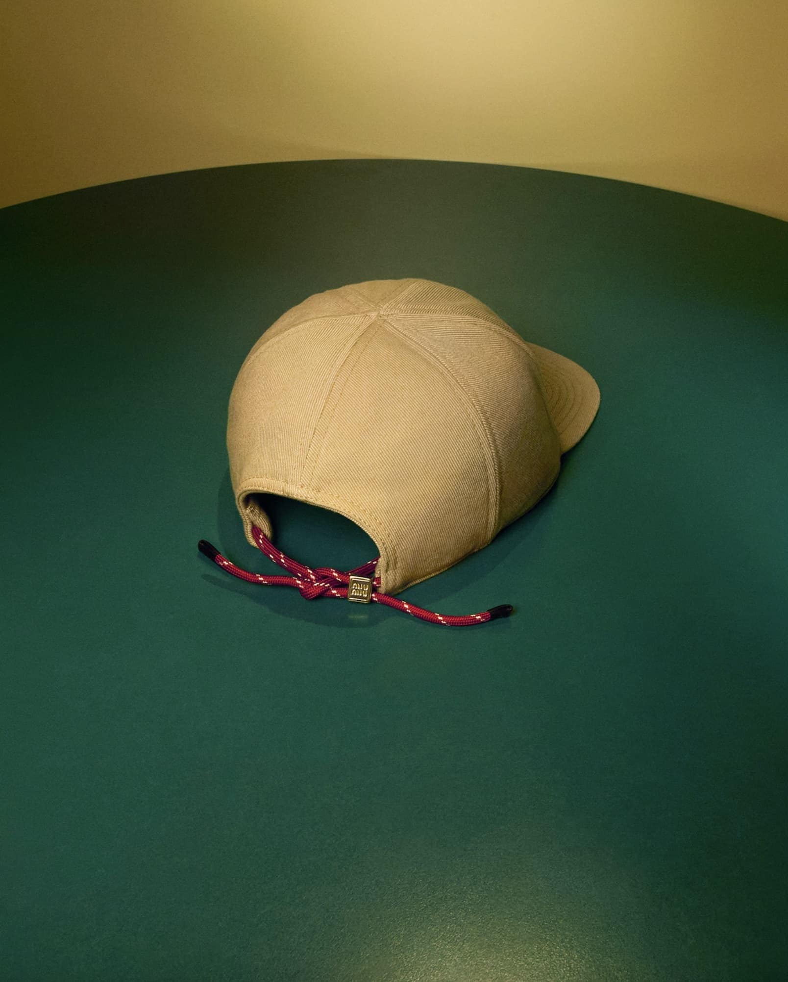 Beige cap resting on a dark green surface with a purple adjustable cord at the back.