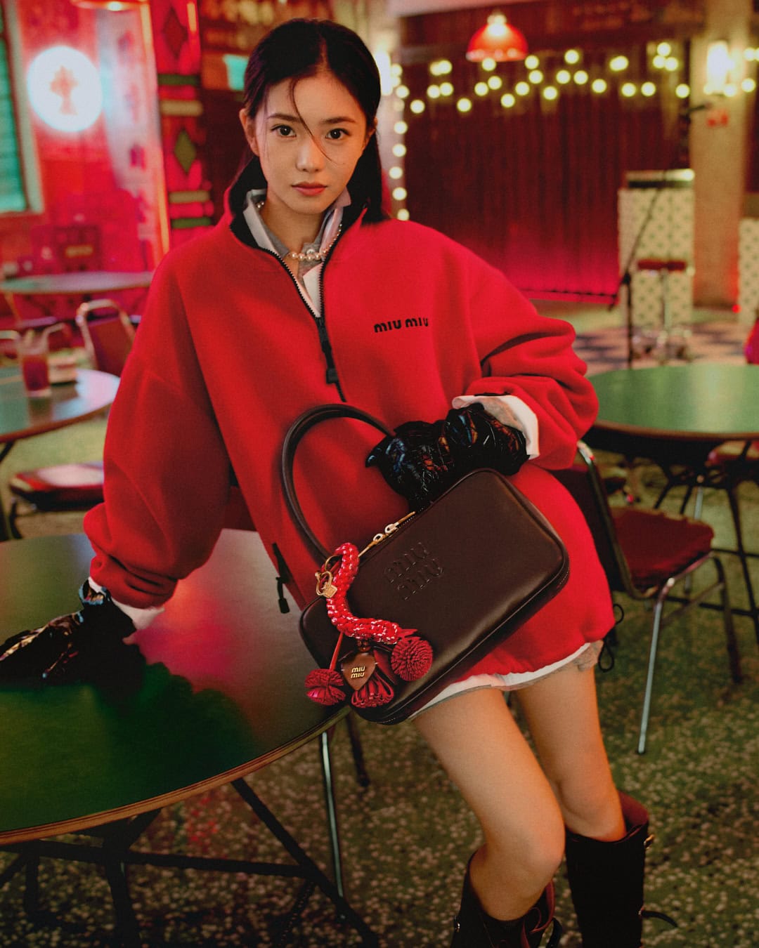 Woman in a red fleece zip-up jacket holding a dark brown handbag while posing indoors at a table.