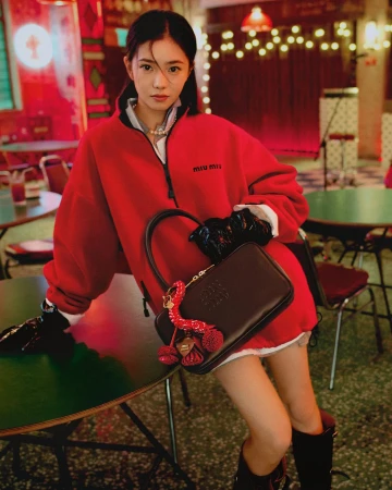 Woman in a red fleece zip-up jacket holding a dark brown handbag while posing indoors at a table.