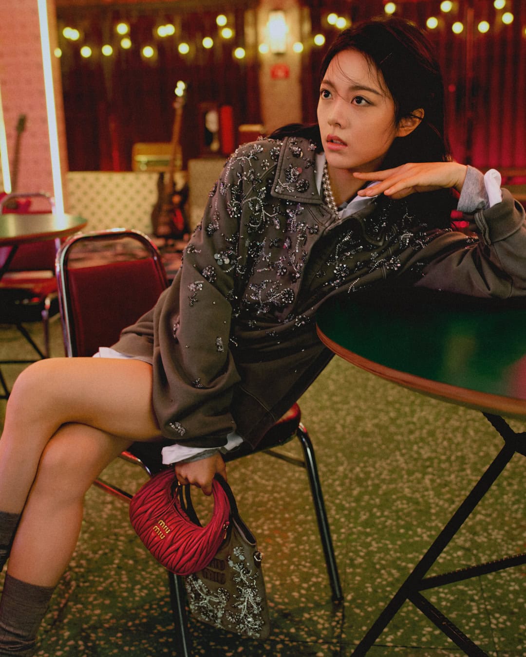 Woman in embellished grey jacket leaning on a table holding a small red handbag in an interior setting.