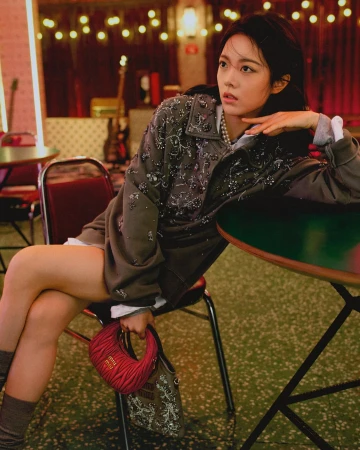 Woman in embellished grey jacket leaning on a table holding a small red handbag in an interior setting.