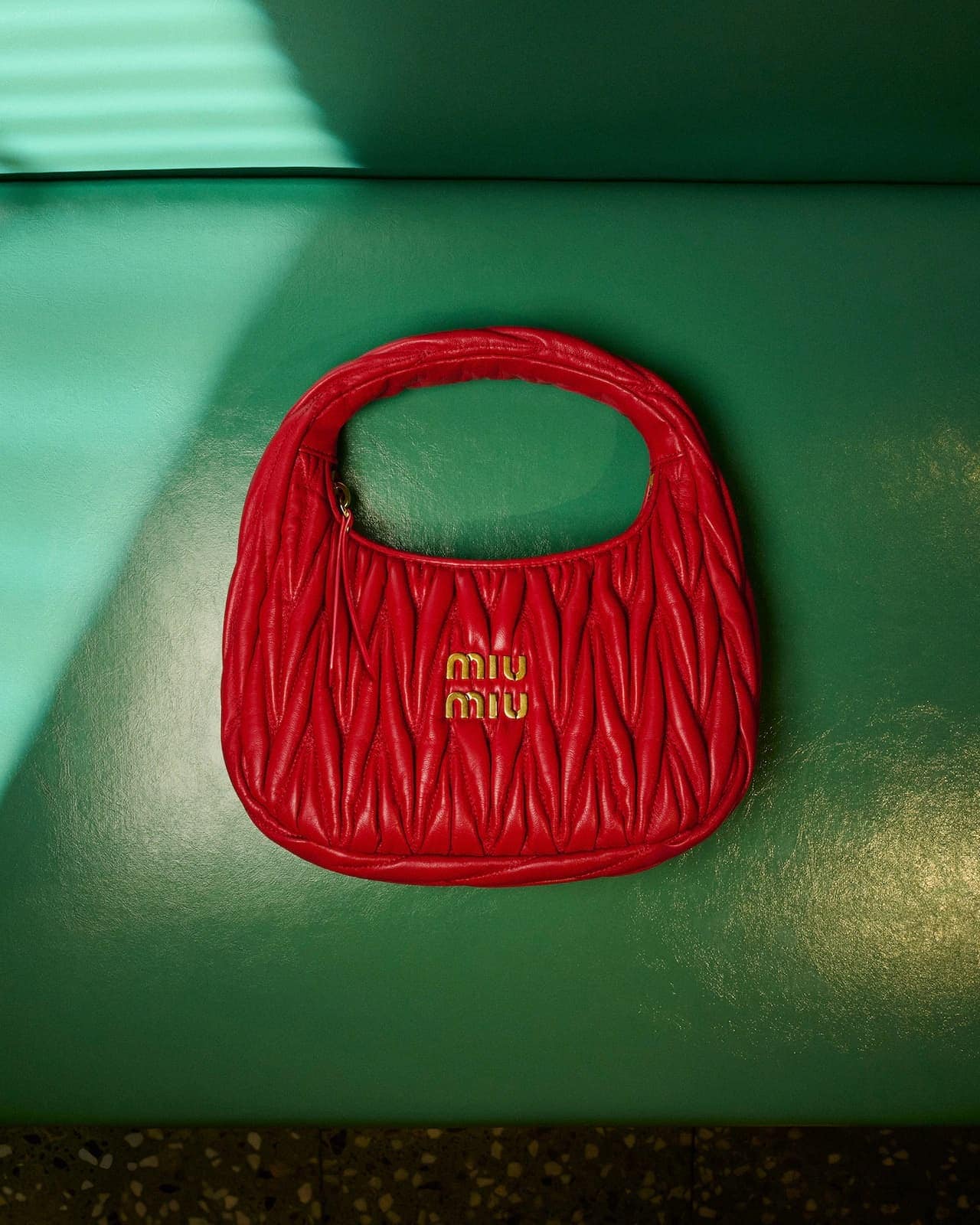 Red quilted leather handbag with gold hardware centered against a smooth green leather background with natural light slanting across