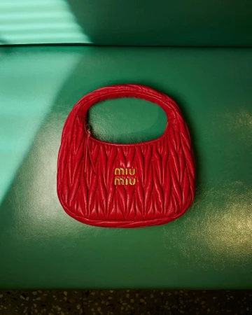 Red quilted leather handbag with gold hardware centered against a smooth green leather background with natural light slanting across