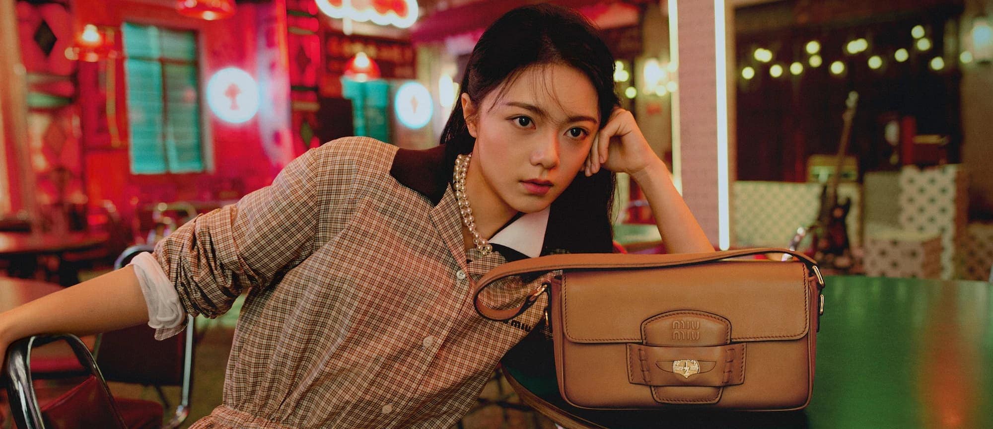 Young woman leaning on a table with a brown leather handbag in a warmly lit interior setting.