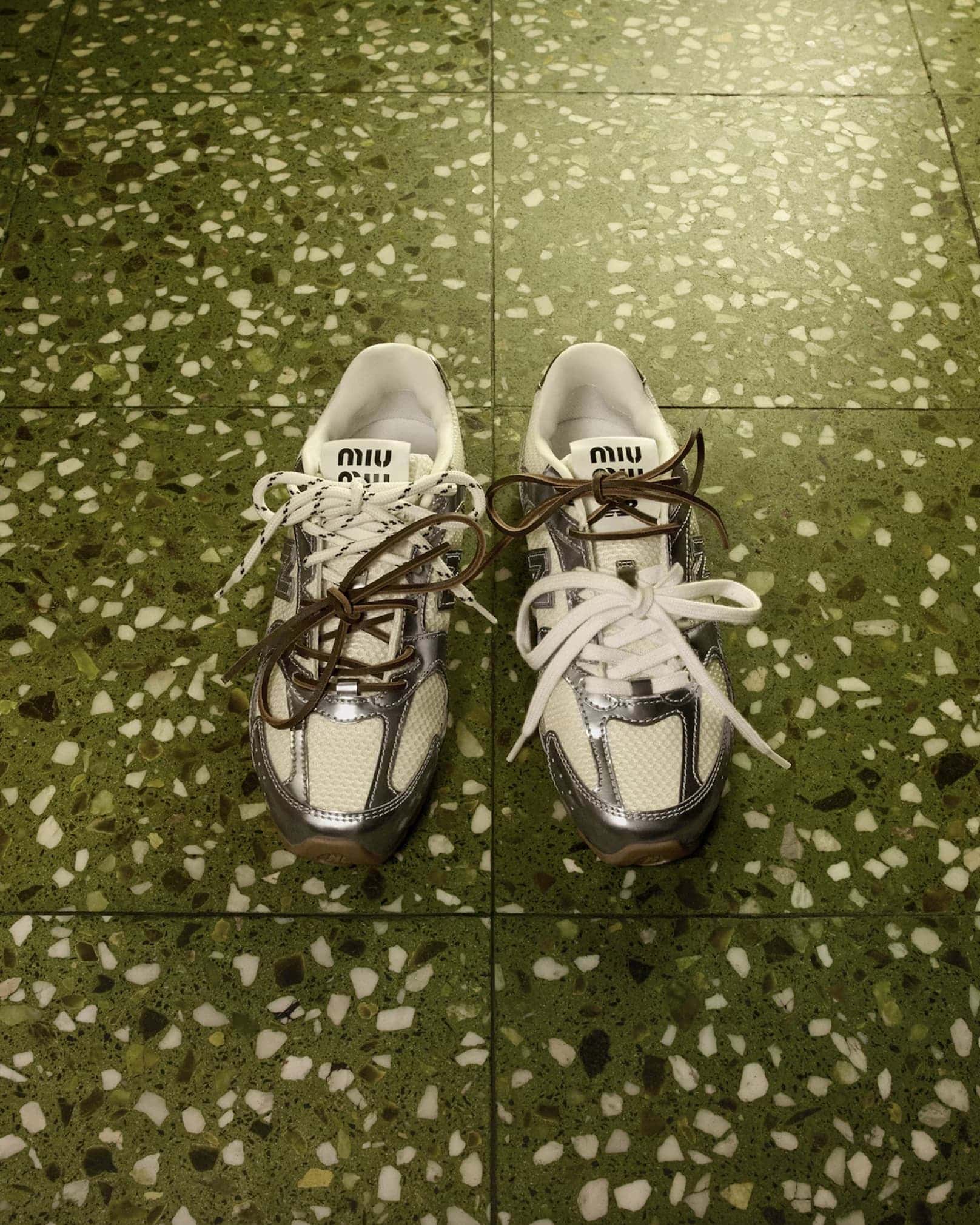 Top-down view of a pair of Miu Miu sneakers with mixed white and brown laces resting on a green terrazzo floor.