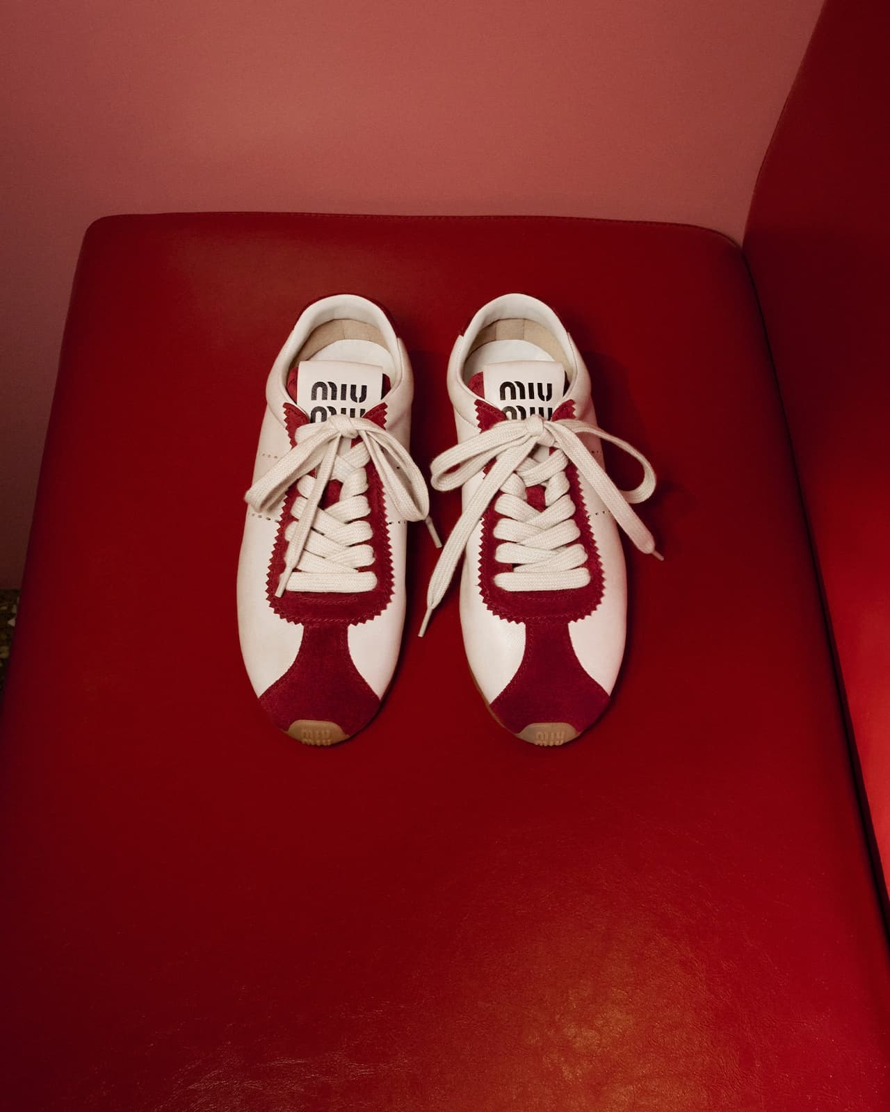 A pair of white and red leather sneakers with wide white laces placed on a red leather surface.