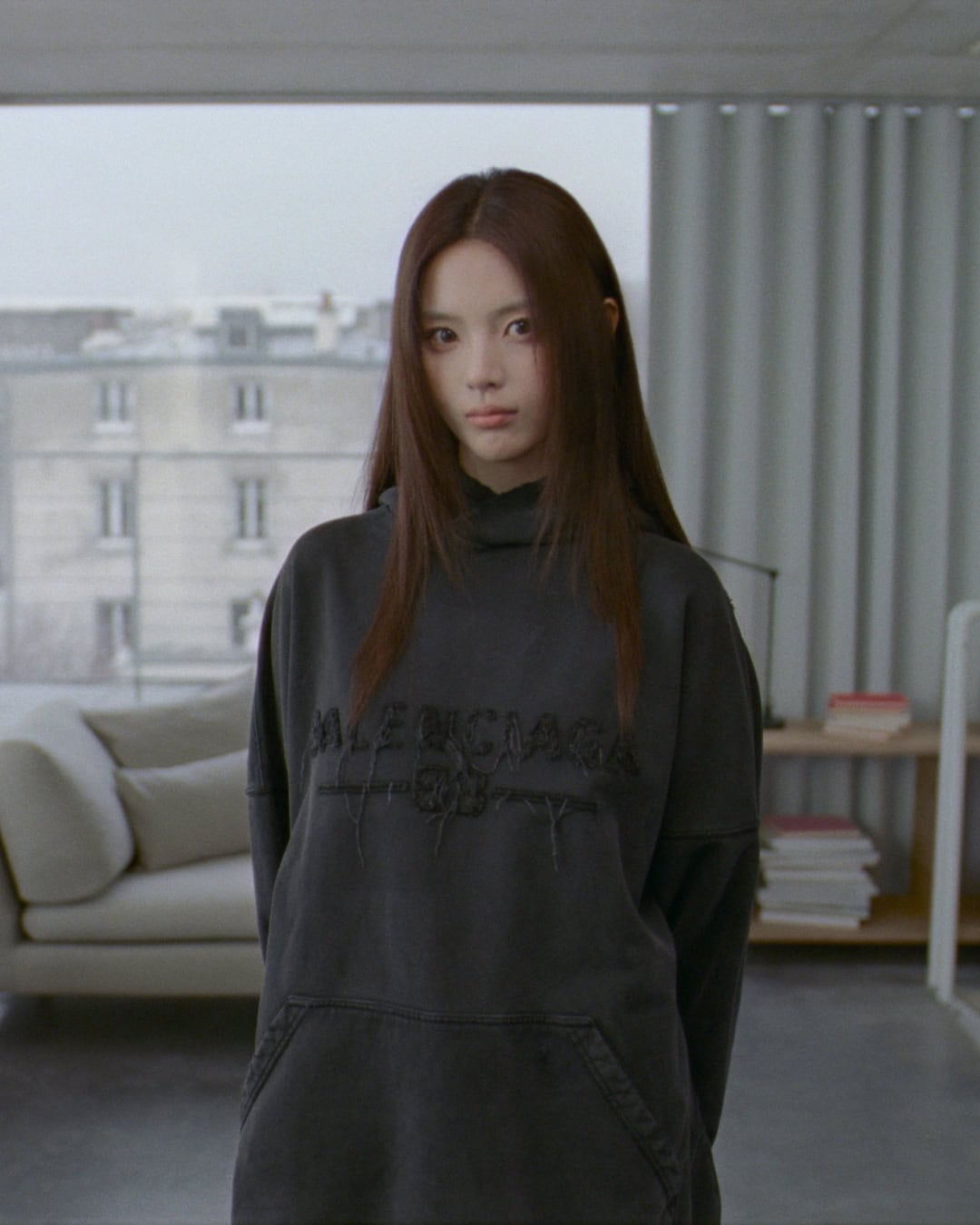 A woman wearing a dark oversized hoodie stands inside a room with a window showing a building exterior in the background.