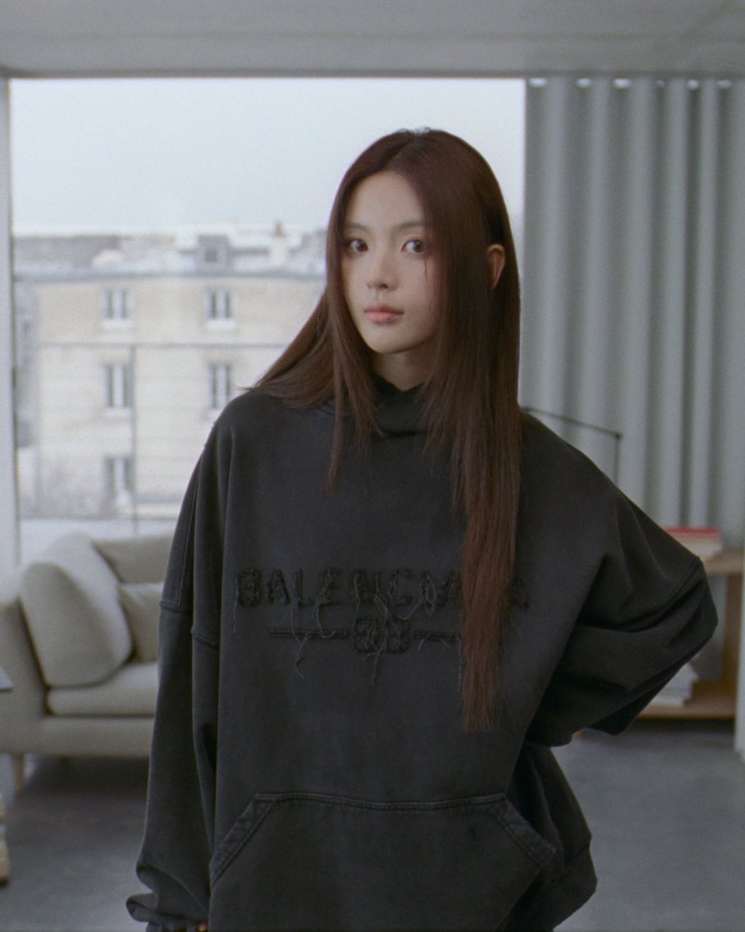 Young woman with long brown hair wearing a dark oversized hoodie standing indoors near a window overlooking a city street.