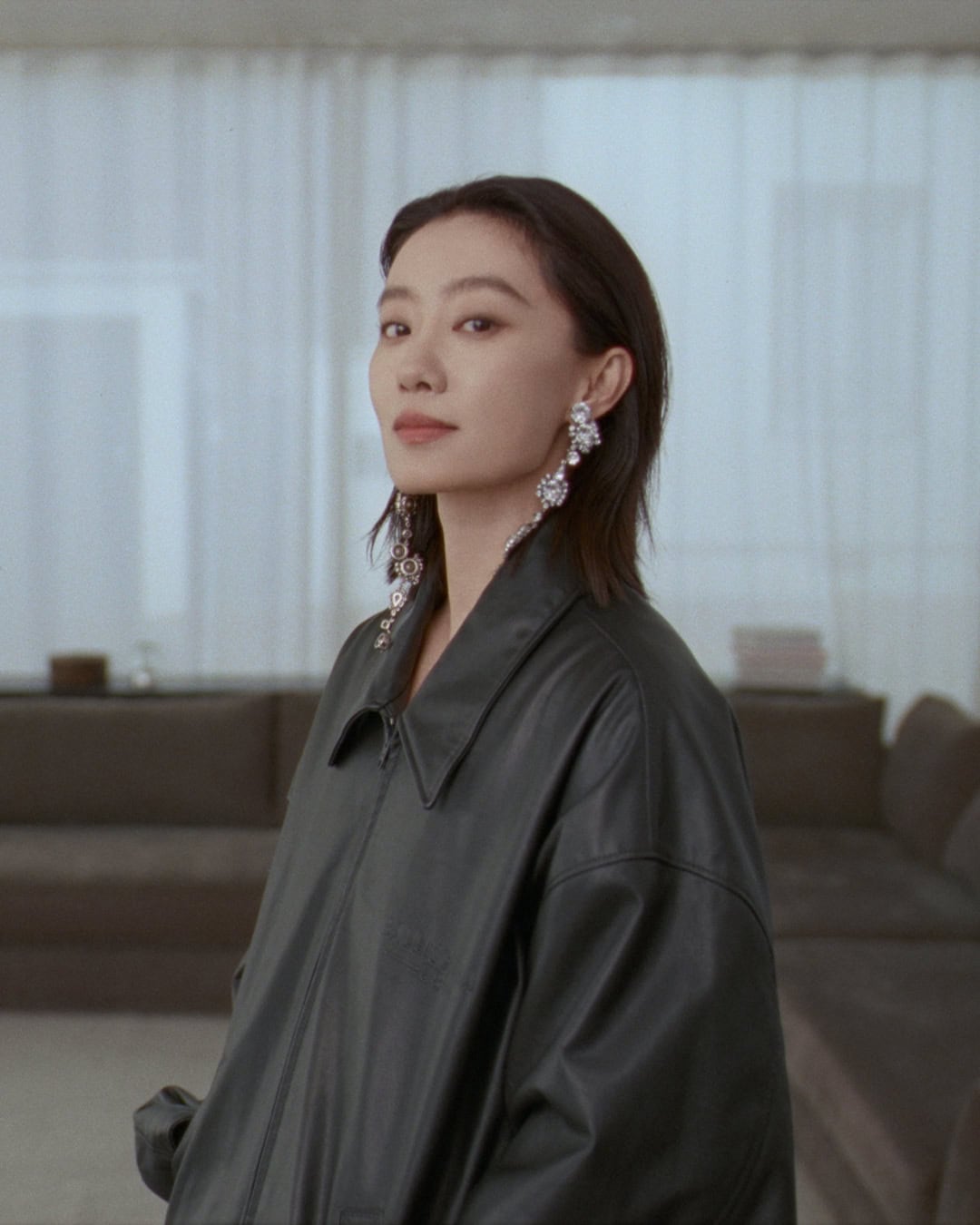 Model wearing dark leather jacket and crystal earrings against domestic interior background