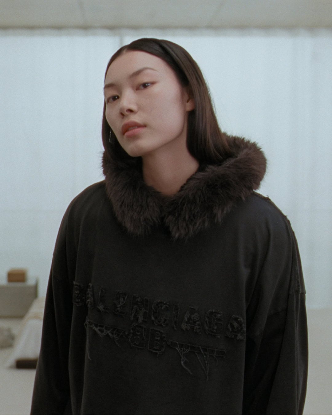 A woman wearing a dark oversized hoodie with a furry collar against a pale seamless background for Balenciaga.