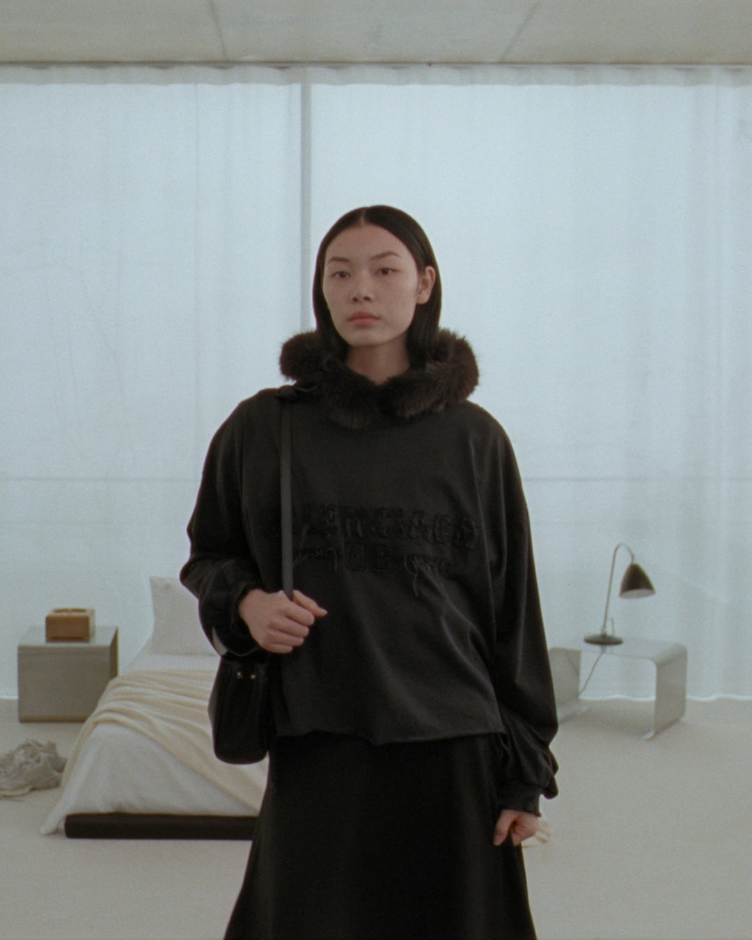 Model wearing a black oversized sweatshirt with a furry collar and holding a dark shoulder bag indoors.