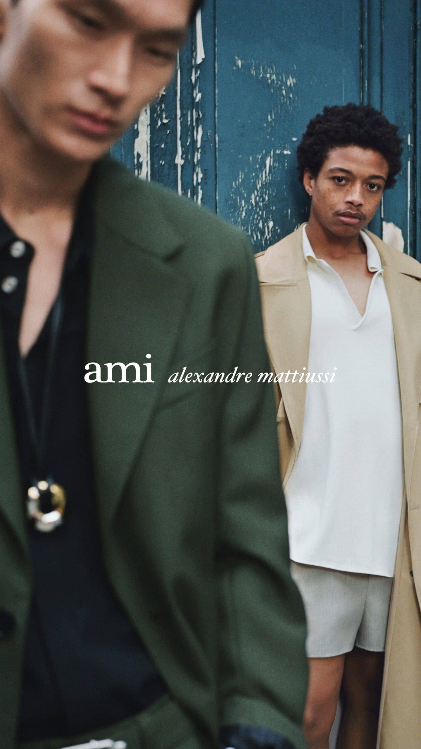 Two male models posing outdoors against a weathered dark blue door for Ami Paris spring 2025 ad campaign.