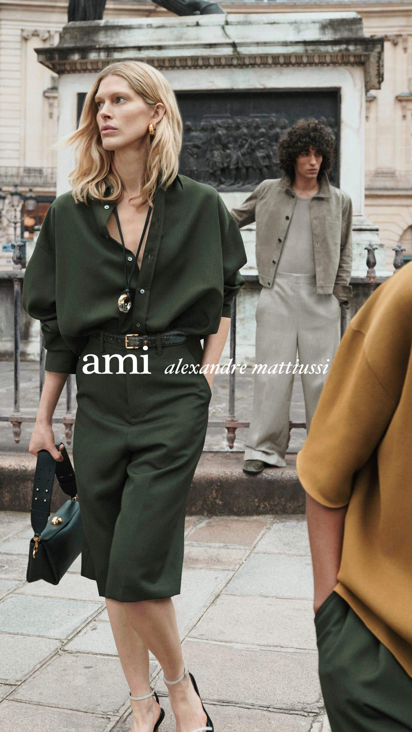 Blonde female model in dark green shirt and trousers holding handbag with other models in background