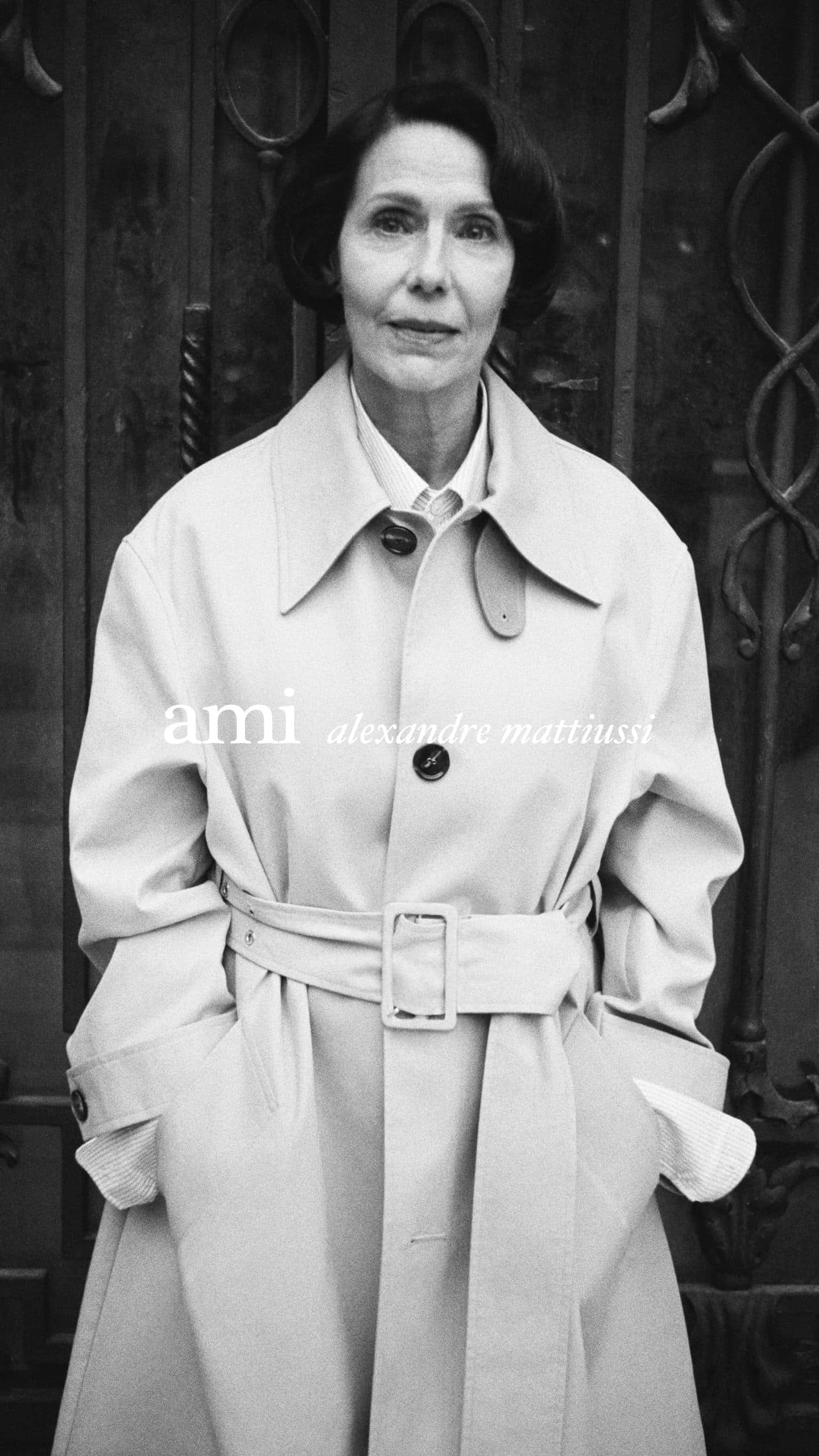 Monochrome portrait of an elderly woman wearing a belted trench coat against a dark wrought iron gate