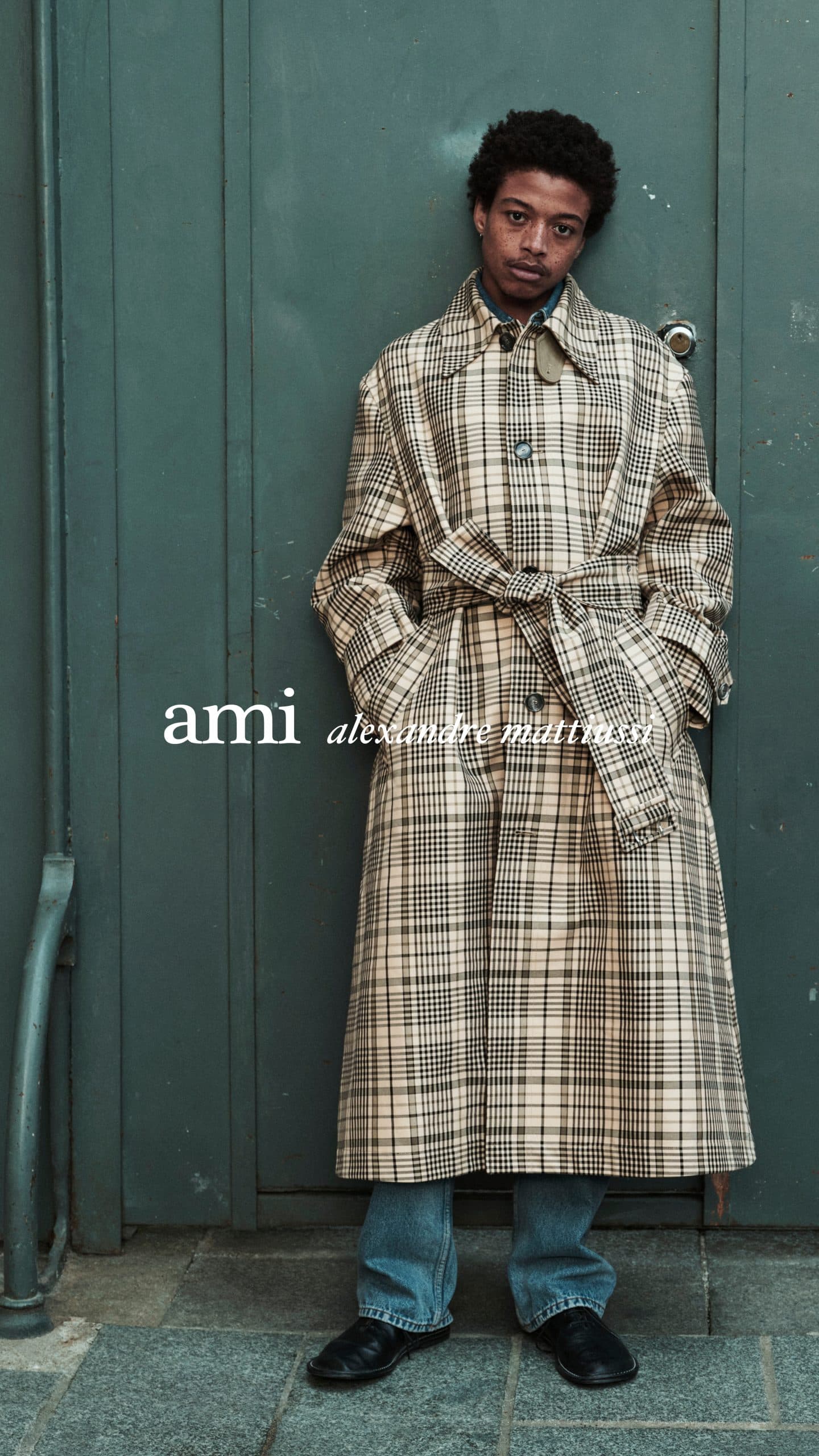 Model wearing a long plaid trench coat leaning against a dark green door for Ami Paris Spring 2025 campaign.