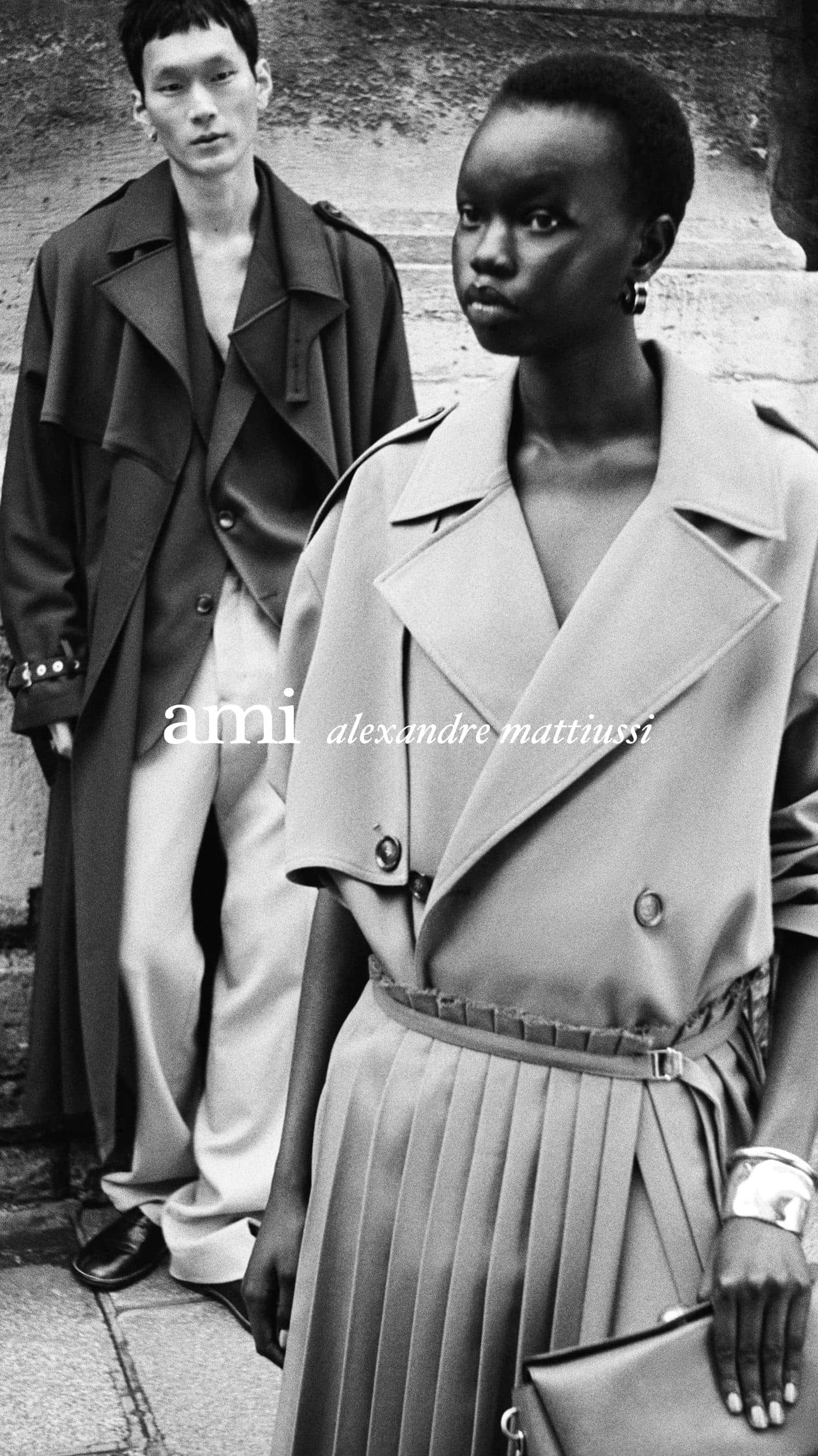 Black and white fashion campaign image featuring two models wearing tailored coats outdoors against a stone architectural backdrop