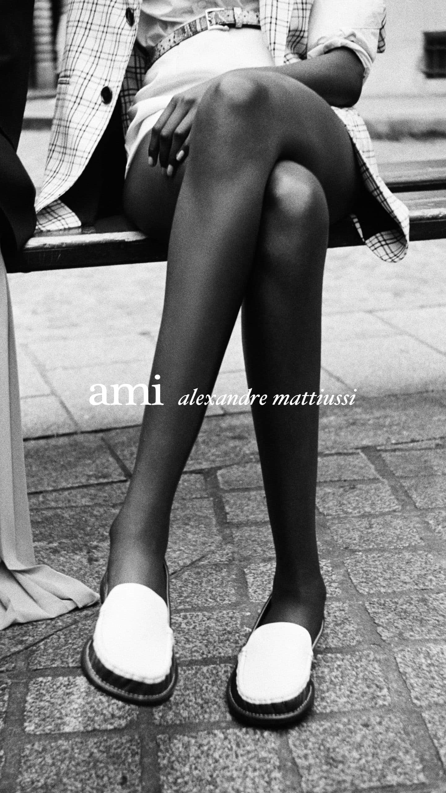 Black and white editorial photo showing a person sitting on a bench wearing loafers, plaid blazer, and shorts, with Ami branding.