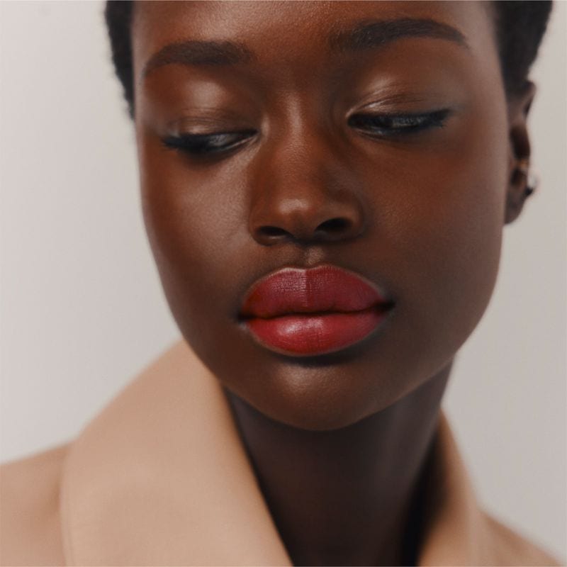 Close-up portrait of a Black model with bold red lipstick wearing a neutral-toned coat against a plain background