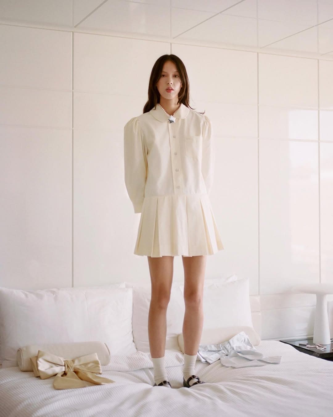 Model standing on a white bed wearing a cream collared dress with pleated skirt and white socks against panelled walls
