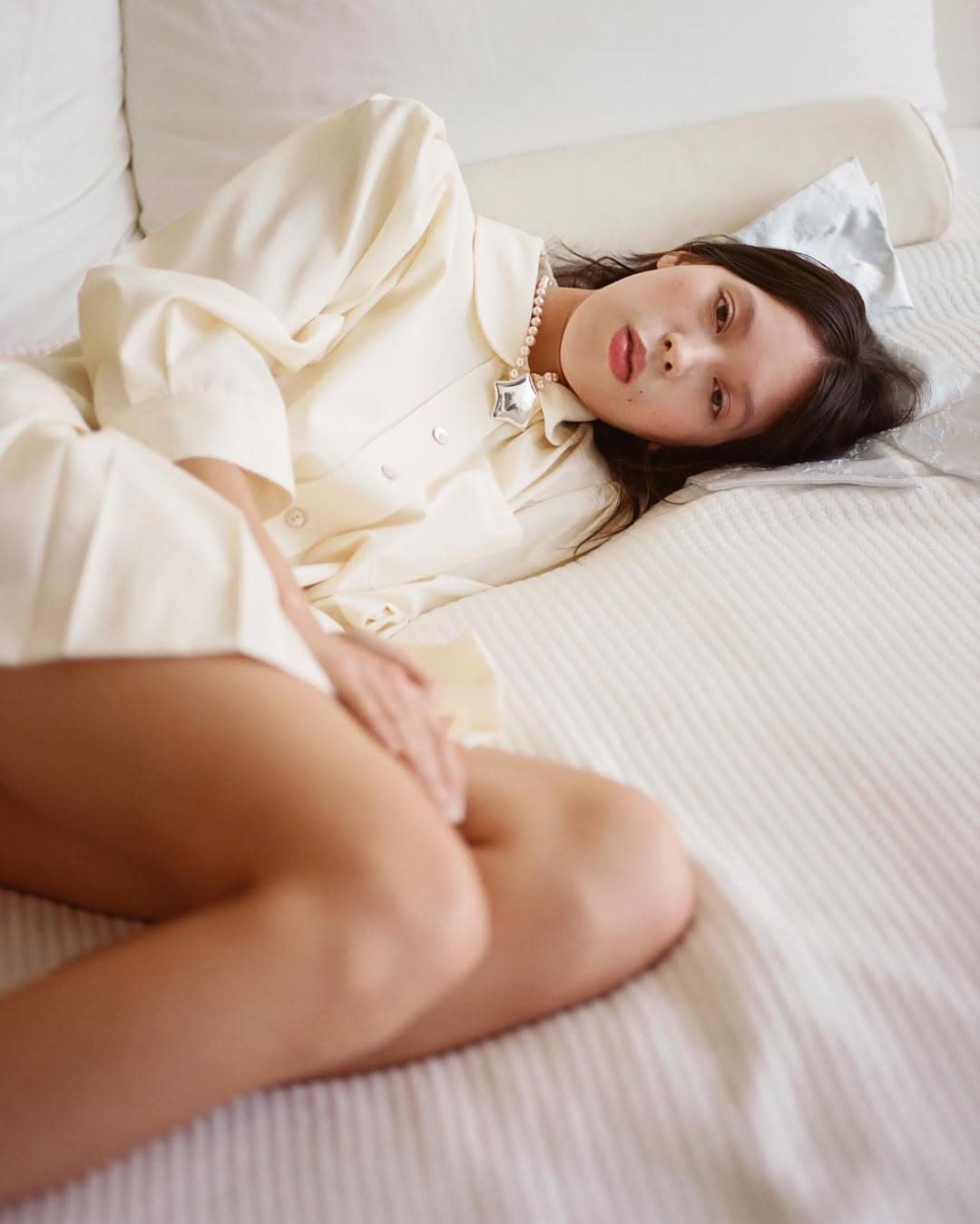Model lying on white hotel bed wearing a cream collared shirt and star necklace for Sandy Liang
