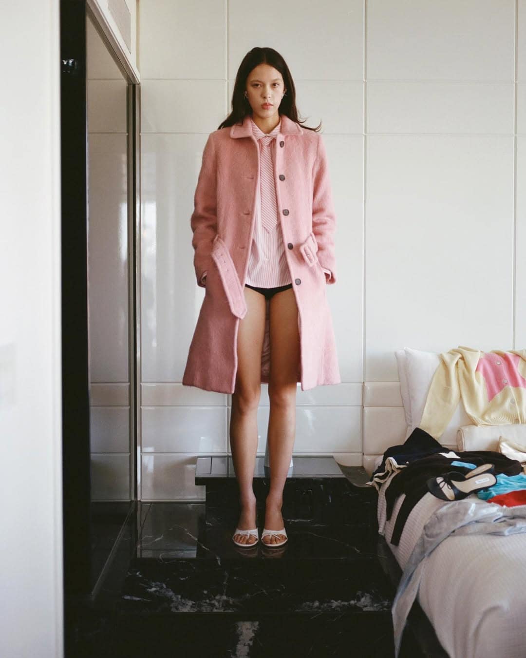 Model standing in a hotel room wearing a pink coat and pink button-down shirt for Sandy Liang resort campaign