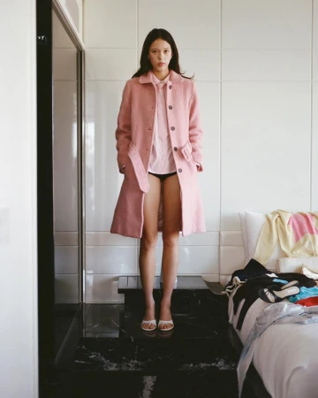 Model standing in a hotel room wearing a pink coat and pink button-down shirt for Sandy Liang resort campaign